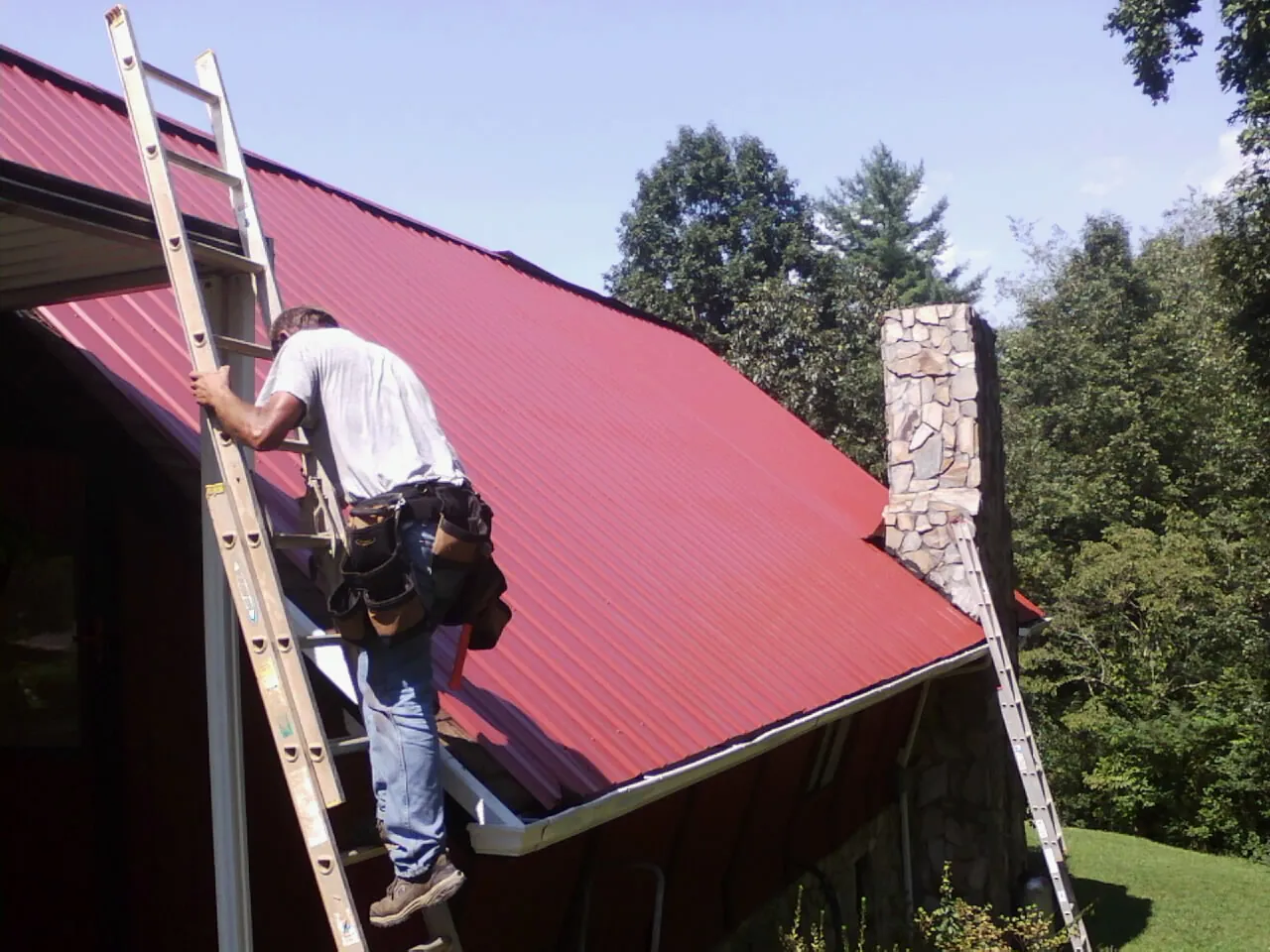 Roofer on a ladder beside a red metal roof and stone chimney — Soffit & Fascia Repair by Roofing Brothers in Stewartville